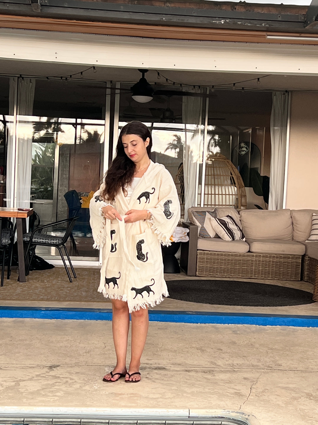 A woman stands on a patio by a pool, wearing the Black Leopard Hooded Cotton Kimono Robe from turkanhome.com, featuring black cat designs. Behind her are outdoor sofas, chairs, a hanging chair, and sliding glass doors.