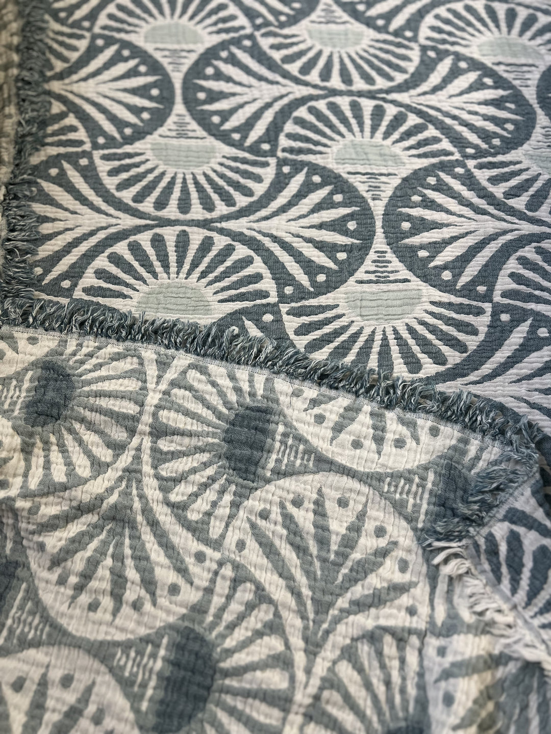 A close-up of the Gingko Muslin Bed Blanket – Teal by turkanhome.com shows blue and white fan-like patterns with radiating lines and dots, a soft textured surface, and frayed edges for a cozy, oversized feel.