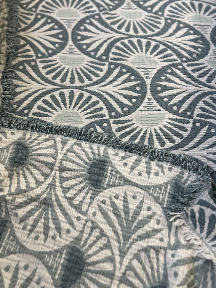 A close-up of the Gingko Muslin Bed Blanket – Teal by turkanhome.com shows blue and white fan-like patterns with radiating lines and dots, a soft textured surface, and frayed edges for a cozy, oversized feel.