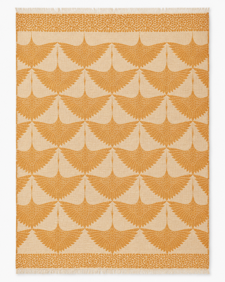 A beige Crane Muslin Throw Blanket from turkanhome.com, featuring a reversible design with a natural border and adorned with symmetrical, orange fan-shaped motifs and an orange floral accent along the top and bottom edges, echoing intricate Turkish-inspired patterns.