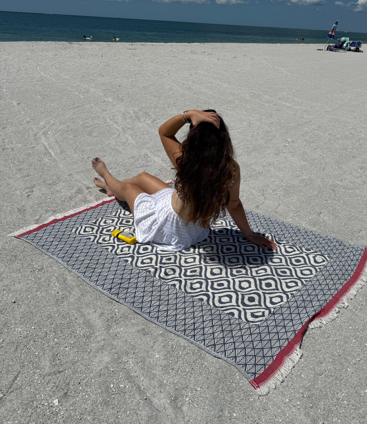 A woman in a white dress sits on the Ikat Throw by turkanhome.com, its reversible geometric pattern facing the sunlit ocean. With her back to the camera, she runs her hand through her hair as sunscreen rests nearby.