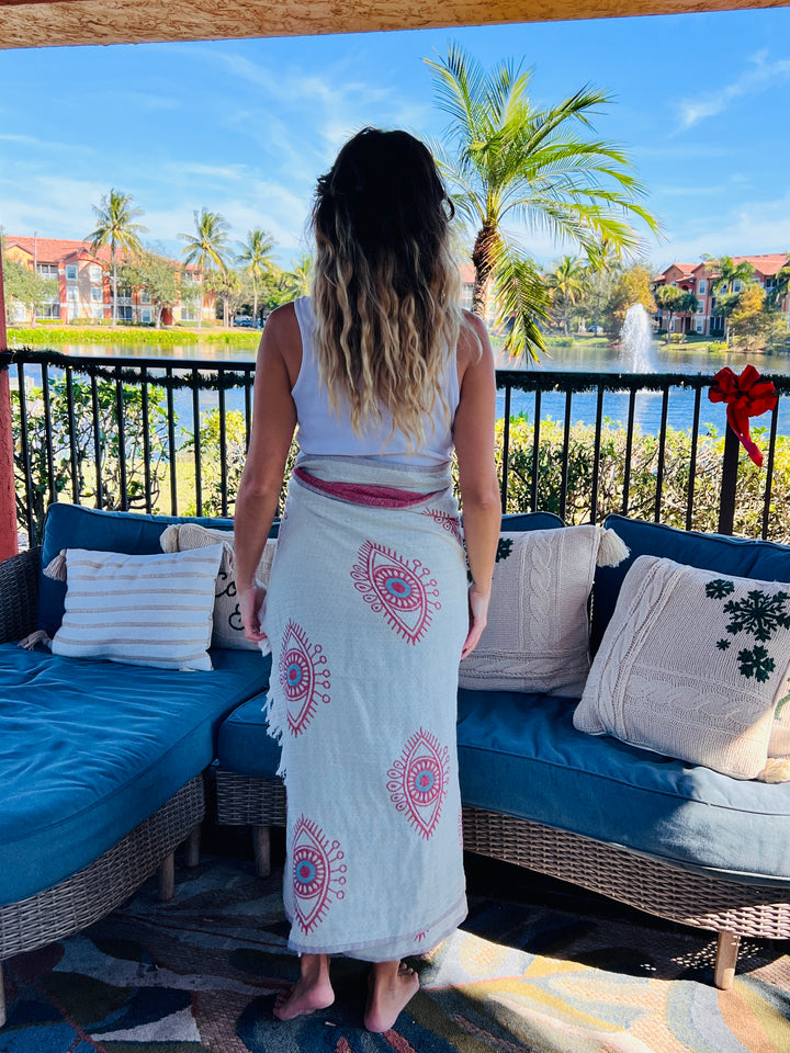 A woman with long, wavy hair stands barefoot on a patio by a palm-lined lake, draped in the Eye Sand Resistant Turkish Beach Towel from turkanhome.com. Cushioned outdoor seating and festive decor complete the relaxing scene.