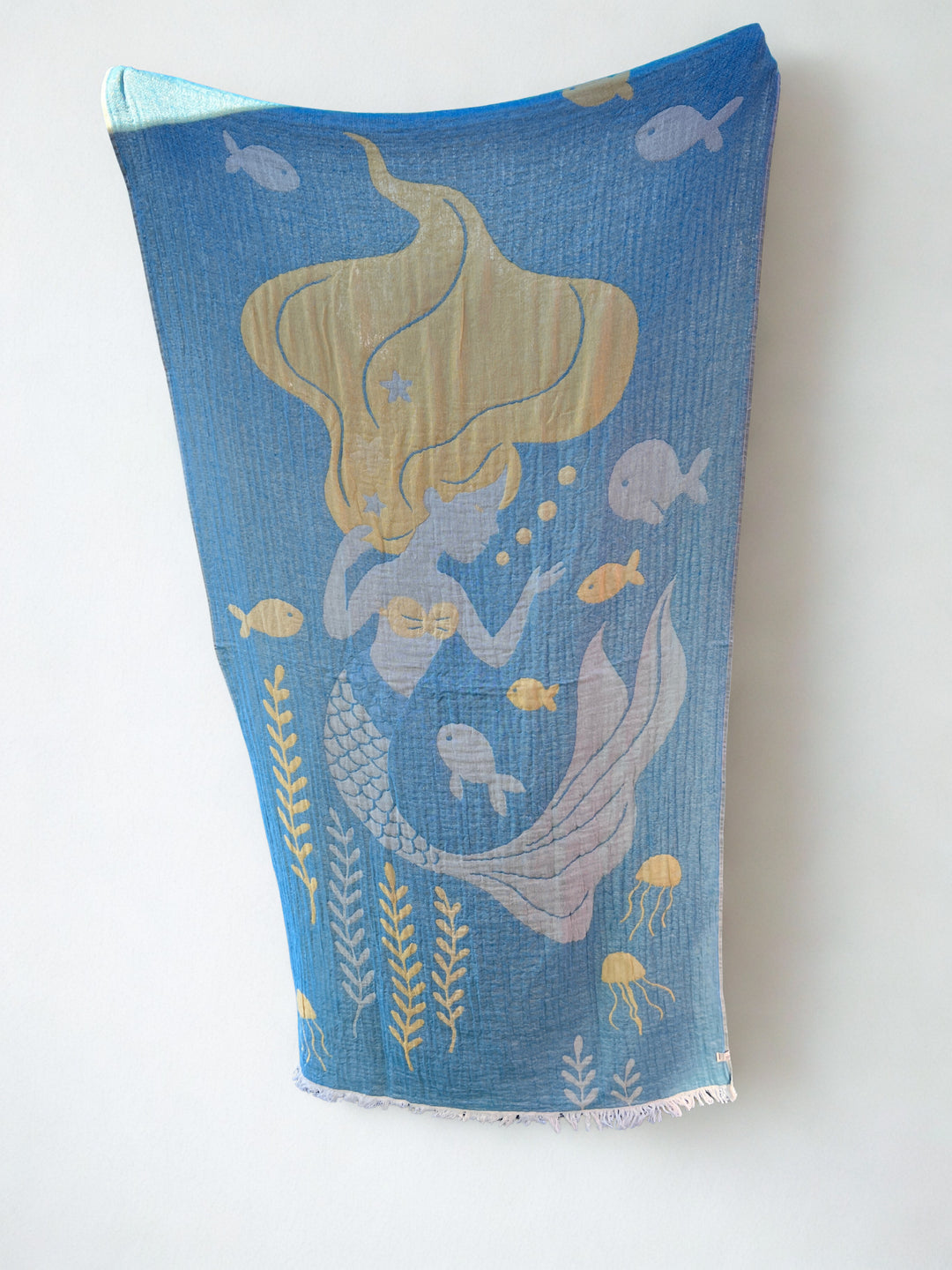 The Mermaid Turkish Towel by turkanhome.com is a blue and gold reversible, quick-drying beach towel with a mermaid amid fish, seaweed, and jellyfish on an ocean-themed background.