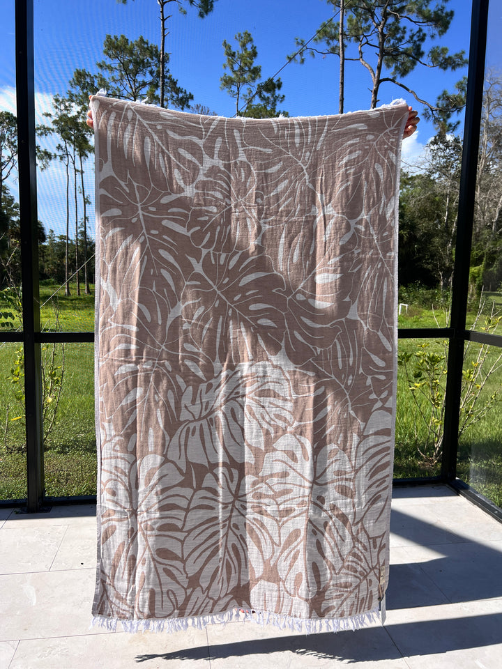 A person holds up the Botanical Turkish Towel from turkanhome.com, featuring a beige and white tropical leaf pattern, outdoors with trees and greenery in the background on a sunny day.