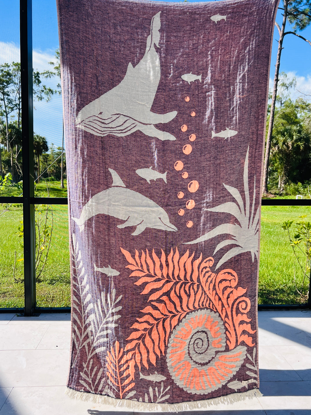 The Dolphin and Whale Turkish Towel from turkanhome.com, made of 100% cotton, features jacquard-woven dolphins, whales, and tropical plants in purple, light gray, and orange hues. It’s pictured outdoors with green grass and trees in the background.