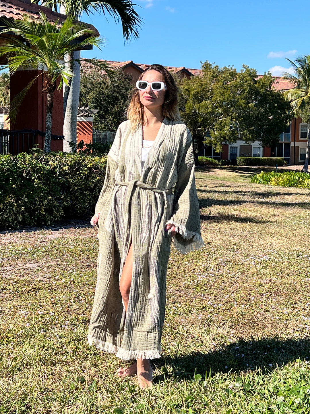 A woman in the "Cactus Robe" by turkanhome.com, perfect for lounge or beach wear, strolls barefoot on grass under the sun, wearing white sunglasses with palm trees and buildings in the background.