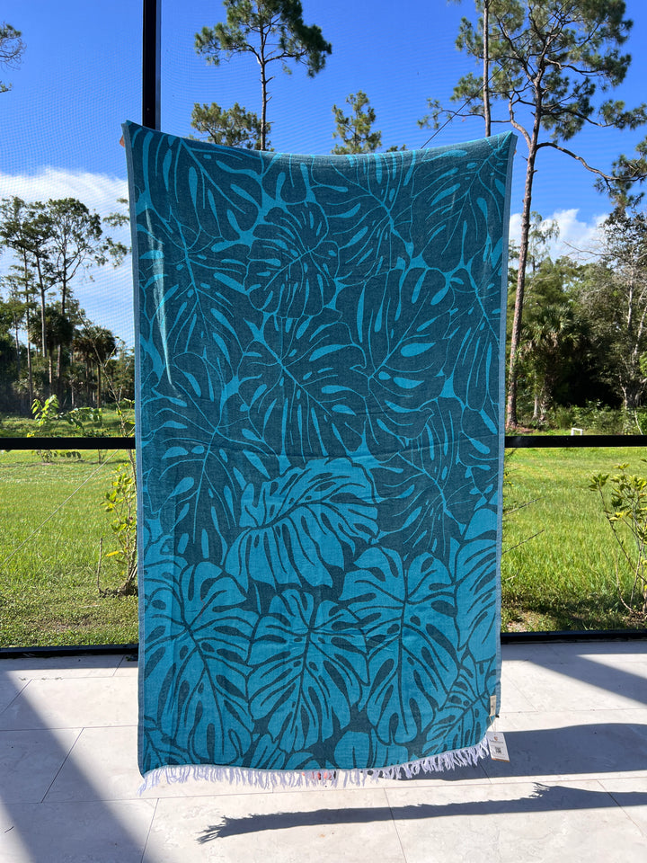 The Botanical Turkish Towel from turkanhome.com, featuring a bold tropical leaf pattern in blue and teal, hangs outdoors in sunlight with lush grass, trees, and a vibrant sky behind it. Made from 100% cotton.