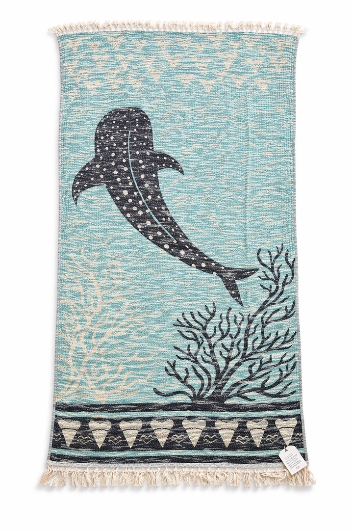 Introducing the Whale Shark Towel – Reversible Turkish Beach Towel from turkanhome.com: a blue woven Turkish cotton towel adorned with a bold black whale shark, detailed coral illustrations, and refined geometric borders. This elegant beach accessory is finished with white fringes on both ends and features a small white tag in the bottom right corner.