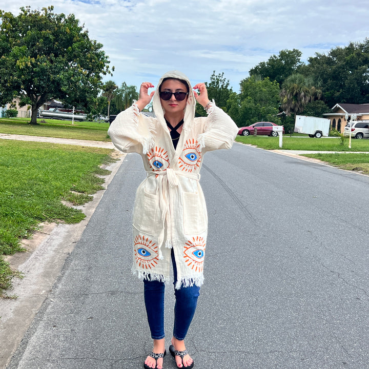 A person stands on a suburban street wearing sunglasses, jeans, sandals, and the Mystic Eye Kimono Robe Short with Pockets and Hood by turkanhome.com. Cream-colored cotton with bold blue and orange eye designs. Trees, grass, houses, and cars are in the background.