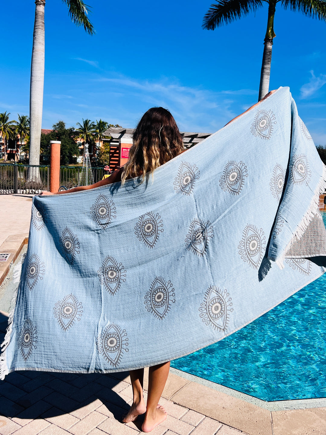 A person stands by a pool, holding the Eye Gray Sand Resistant Turkish Beach Towel from turkanhome.com outstretched behind them. Palm trees and blue sky in the background hint at a sunny, tropical setting.