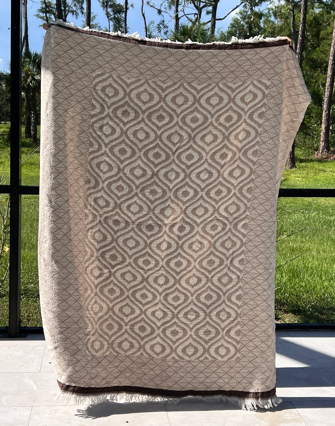 The Ikat Throw from turkanhome.com, featuring a beige and cream reversible geometric pattern, is held upright in front of a window, with views of green grass and tall trees in the background.