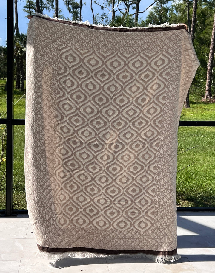 The Ikat Throw from turkanhome.com, featuring a beige and cream reversible geometric pattern, is held upright in front of a window, with views of green grass and tall trees in the background.