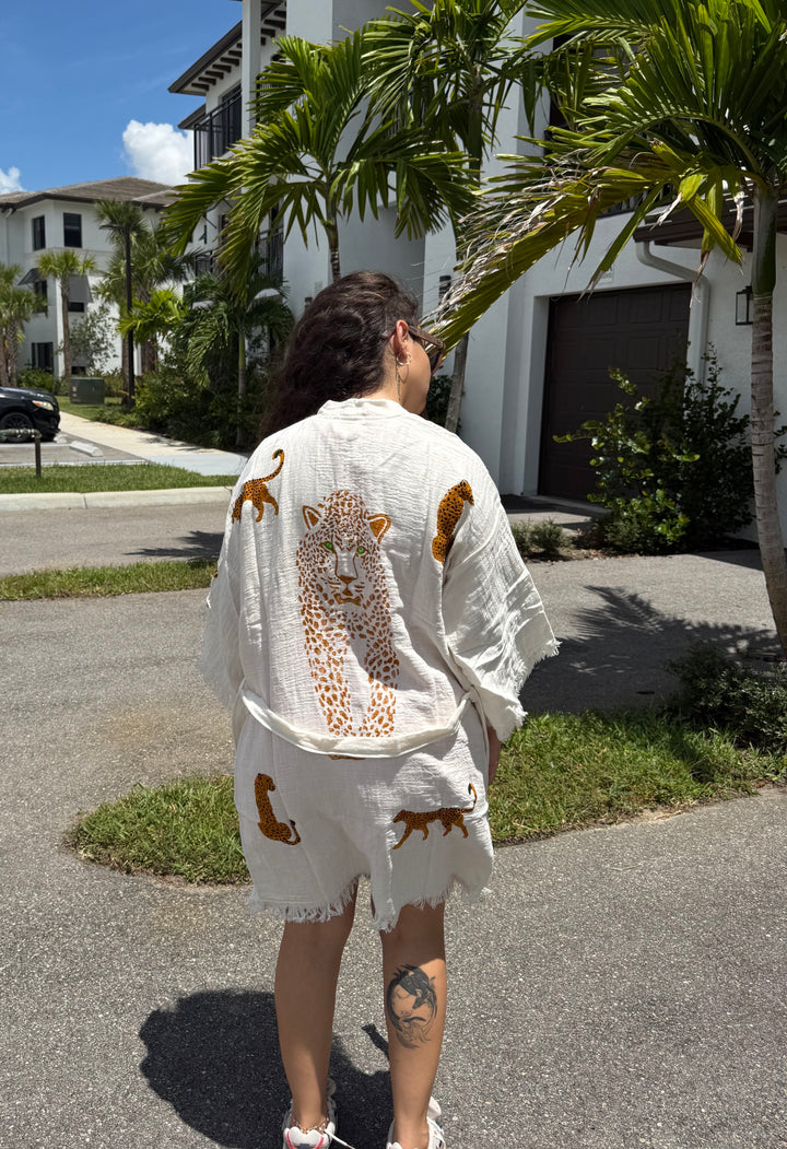 A woman stands outdoors in the sunlight, facing away. She wears the turkanhome.com Leopard Cotton Kimono Robe-Short, featuring a light color and cheetah prints on the back, paired with shorts. A snake tattoo decorates her right calf, while palm trees and houses are visible in the background.