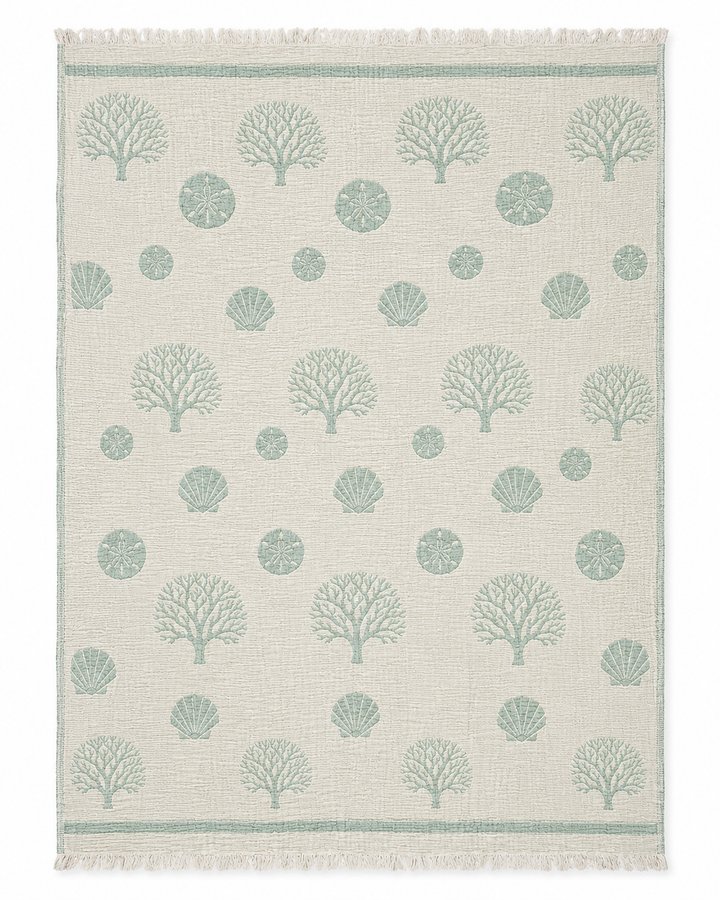 Experience coastal comfort with the Coastal Muslin Throw Blanket – Reversible Turkish Cotton by turkanhome.com, featuring a beige backdrop with green coral, seashell, and sand dollar patterns arranged in rows; fringed edges on the short sides add a classic Turkish touch reminiscent of seaside style.
