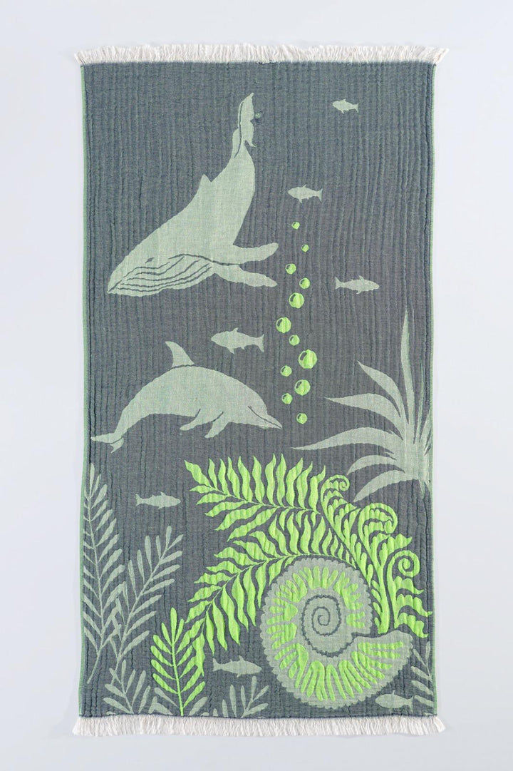 The Dolphin and Whale Turkish Towel by turkanhome.com is a rectangular gray 100% cotton towel, decorated with dolphins, whales, fish, sea plants, bubbles, and a large green seashell pattern plus white fringe on both short edges.