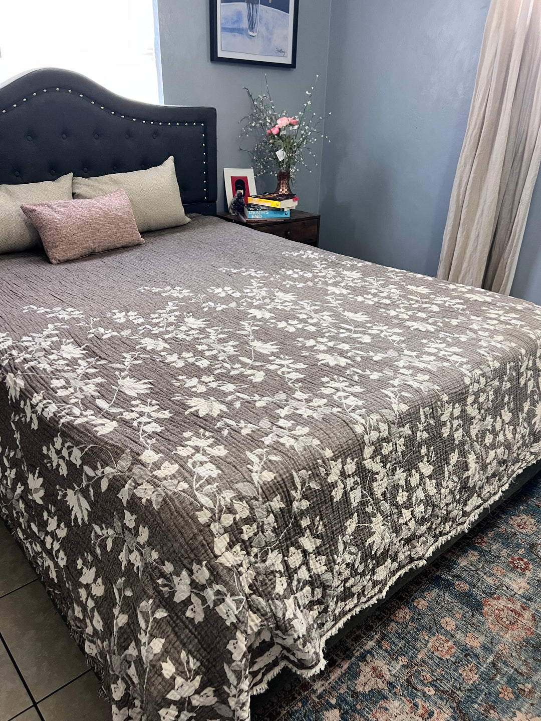 A neatly made bed with a gray and white floral quilt and several pillows, featuring the Bougainvillea Muslin Bed Blanket – Brown (Queen) by turkanhome.com, sits next to a nightstand with books, a framed photo, and flowers in soft light.
