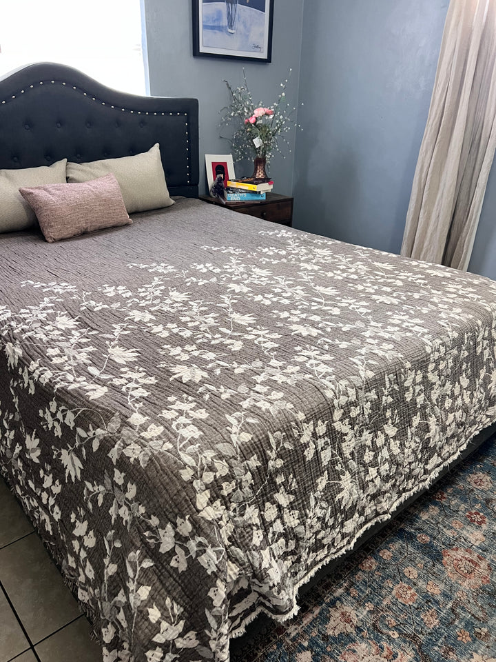 A neatly made bed with a gray and white floral quilt and several pillows, featuring the Bougainvillea Muslin Bed Blanket – Brown (Queen) by turkanhome.com, sits next to a nightstand with books, a framed photo, and flowers in soft light.
