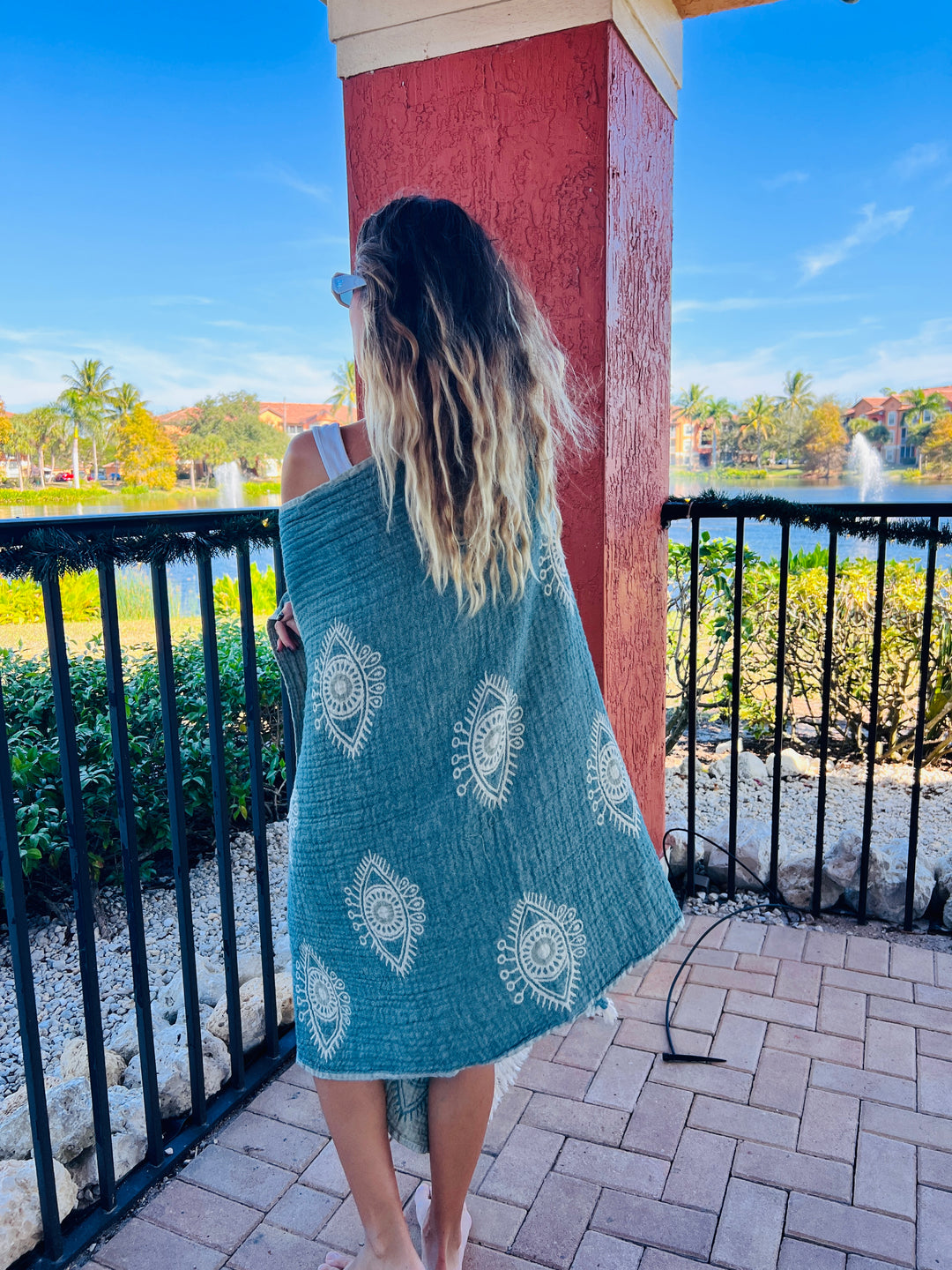 A woman with wavy blonde hair stands barefoot on a patio, facing a lake and palm trees under a sunny sky, wrapped in the Eye Sage Sand Resistant Turkish Beach Towel by turkanhome.com.