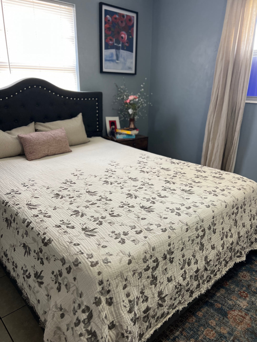 A cozy bedroom features a neatly made bed with the Bougainvillea Muslin Bed Blanket – Brown| Queen from turkanhome.com, decorative pillows, gray-blue walls, framed art above the bed, a nightstand with flowers, and beige-curtained windows letting in light.