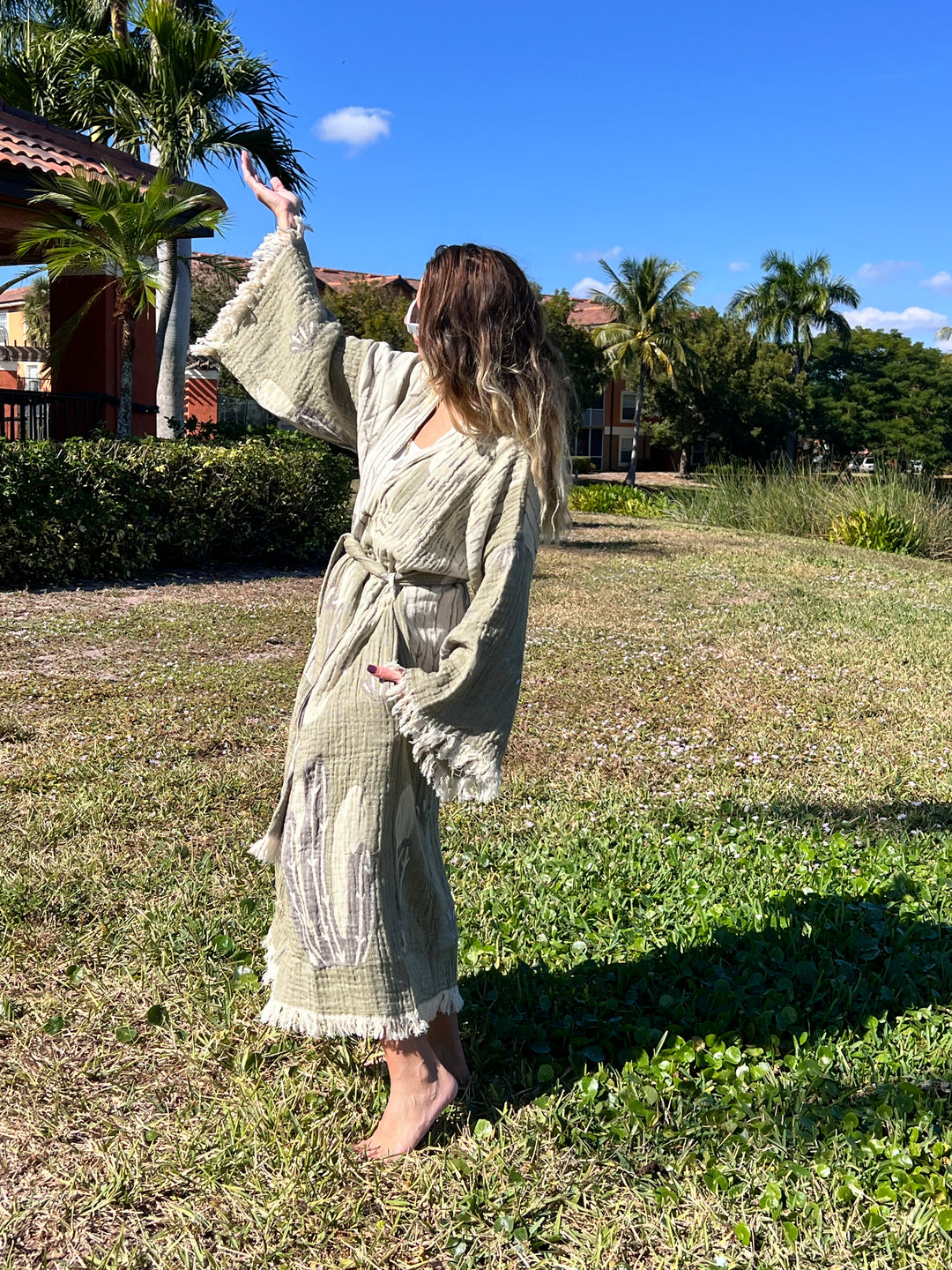 A woman wearing the Cactus Robe by turkanhome.com stands barefoot on grass with one arm raised. Palm trees, bushes, and buildings appear in the sunny background beneath a clear blue sky—ideal lounge or beach wear.