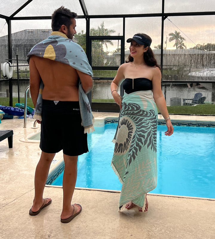 By the pool, a man has a Turkish towel from turkanhome.com draped over his shoulders with black swim shorts, while a smiling woman wears a black swimsuit and wraps herself in the Dolphin and Whale Turkish Towel.