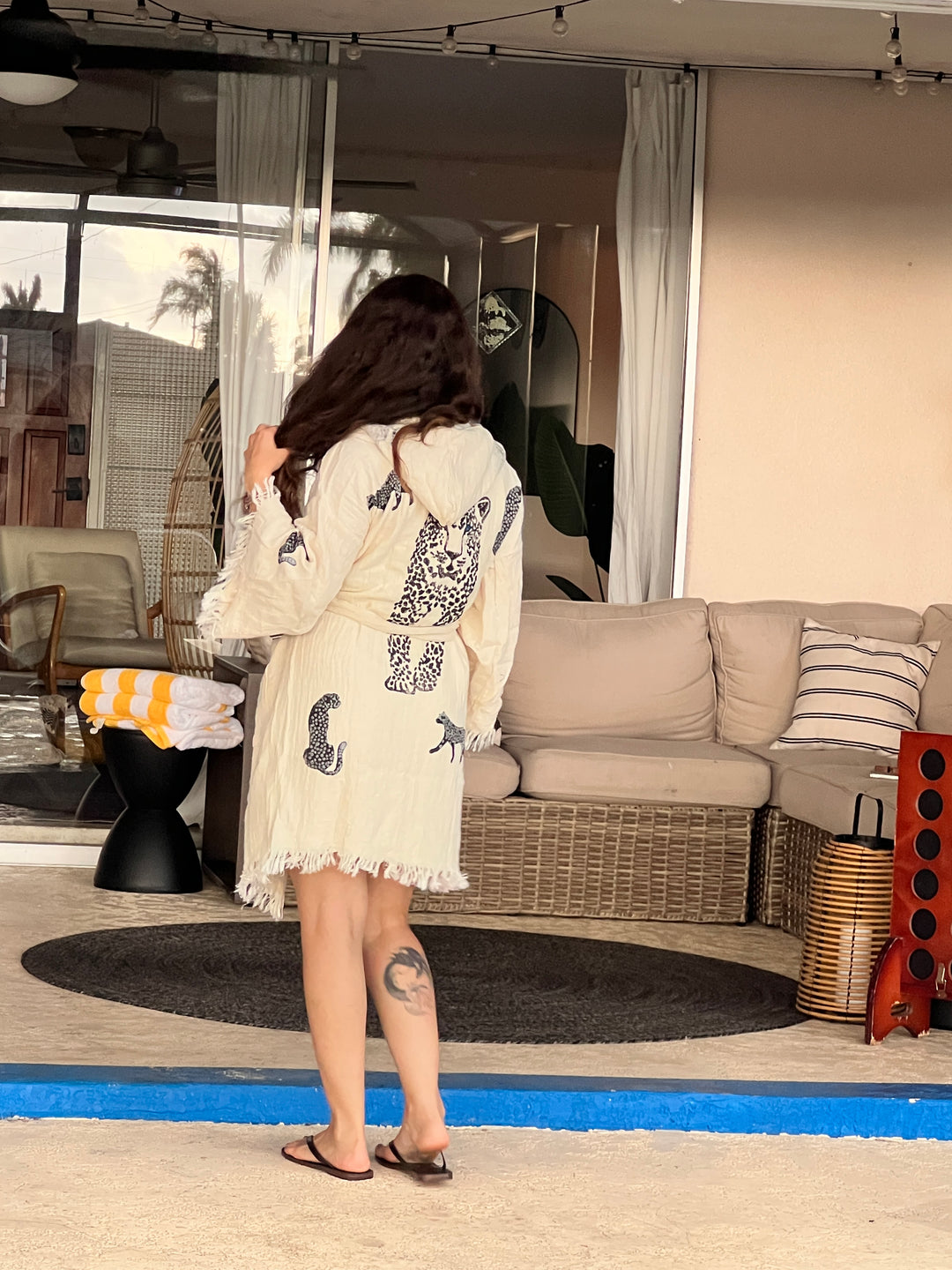 A woman with long brown hair, wearing a Black Leopard Hooded Cotton Kimono Robe by turkanhome.com as a beach cover-up and flip-flops, stands by outdoor wicker furniture with a rolled towel, facing away from the camera.
