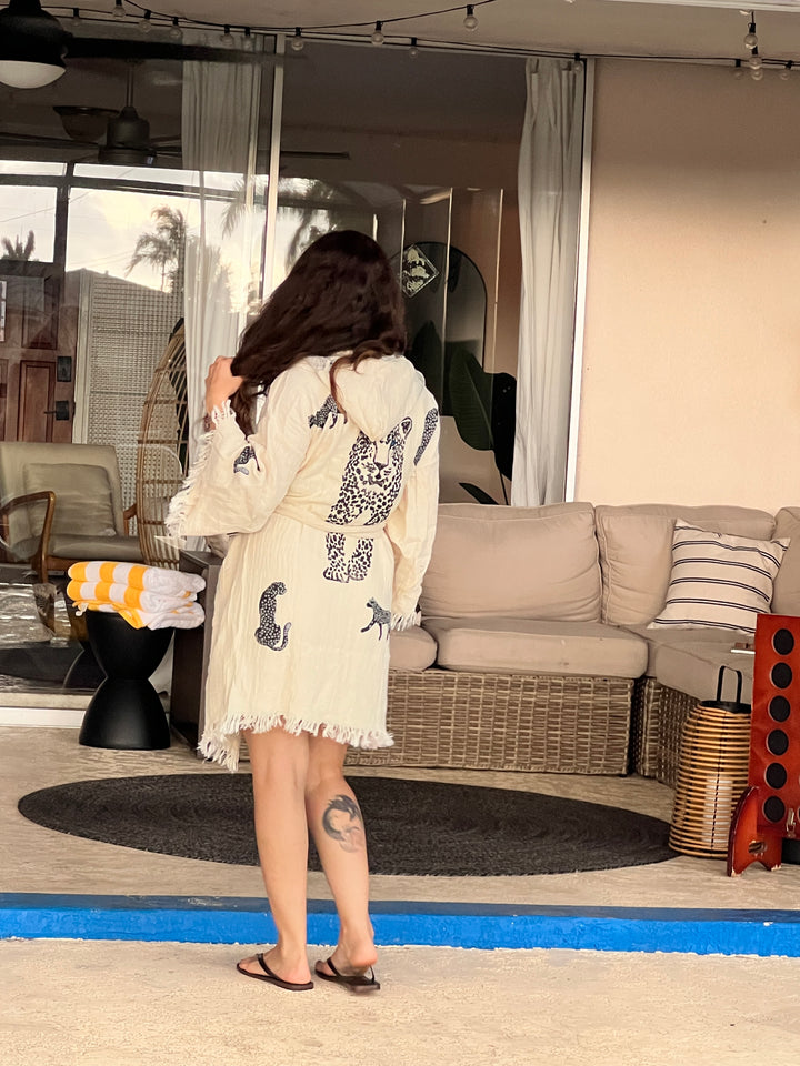 A woman with long brown hair, wearing a Black Leopard Hooded Cotton Kimono Robe by turkanhome.com as a beach cover-up and flip-flops, stands by outdoor wicker furniture with a rolled towel, facing away from the camera.