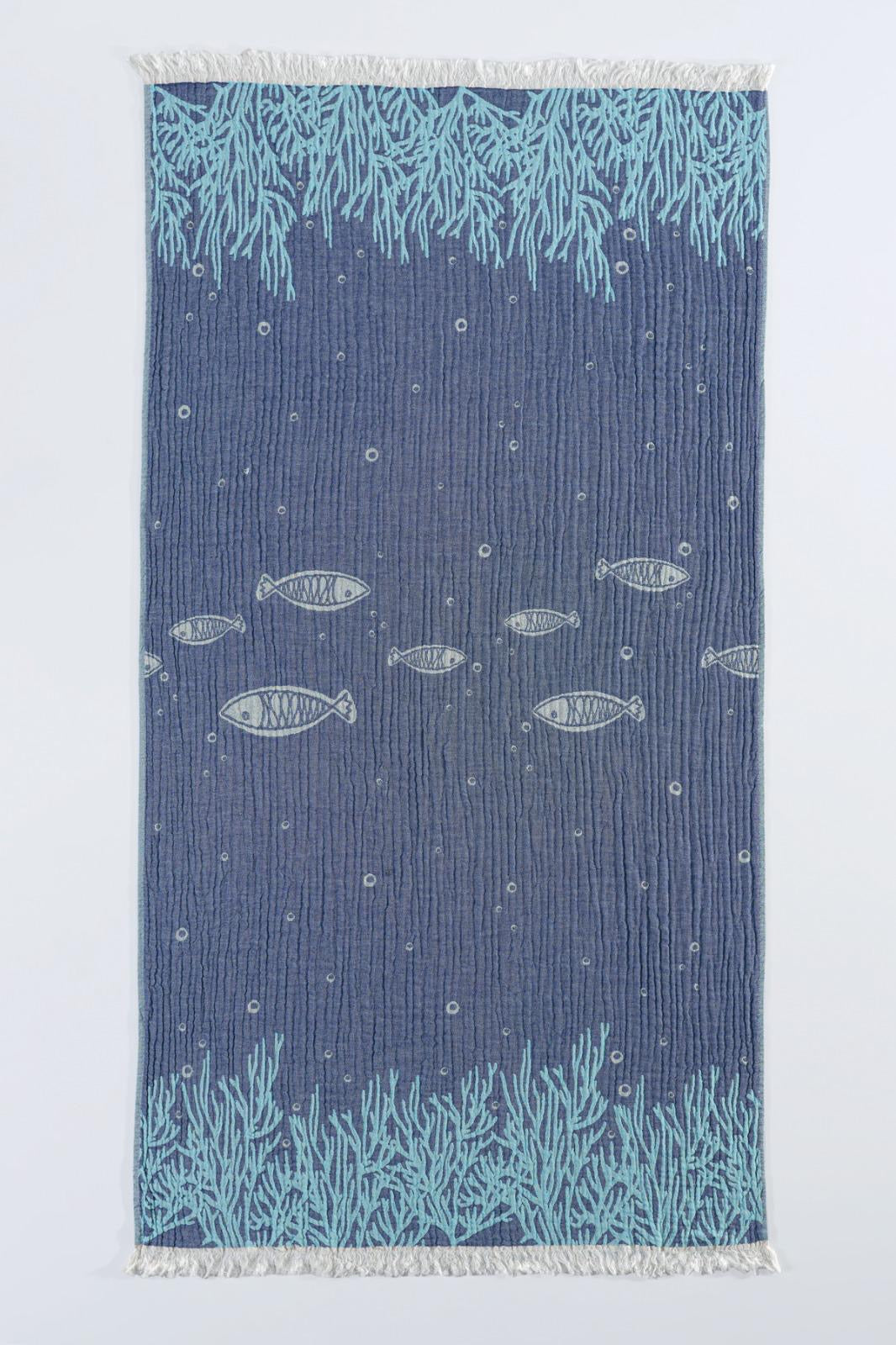 The Fishes and Coral Turkish Towel by turkanhome.com is a rectangular blue cotton jacquard towel with light blue coral at the ends, central fish swimming among bubbles, and white fringe on both shorter sides.