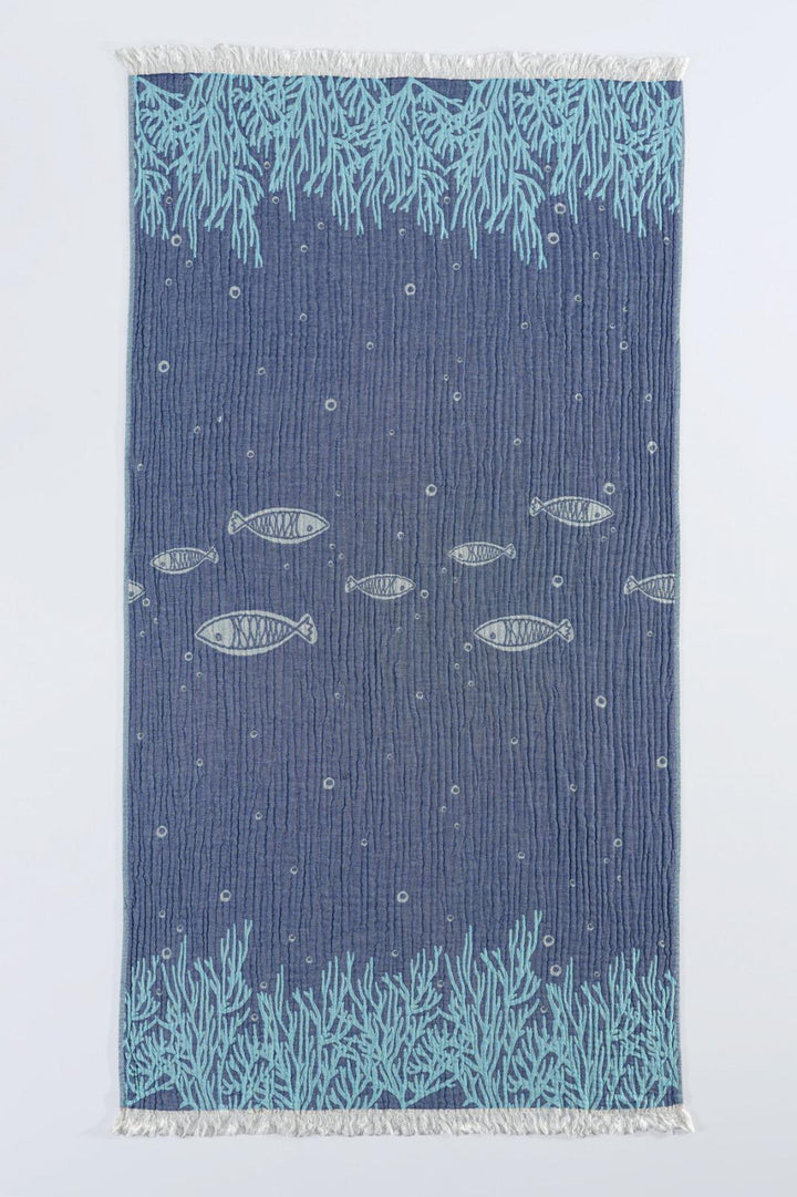 The Fishes and Coral Turkish Towel by turkanhome.com is a rectangular blue cotton jacquard towel with light blue coral at the ends, central fish swimming among bubbles, and white fringe on both shorter sides.