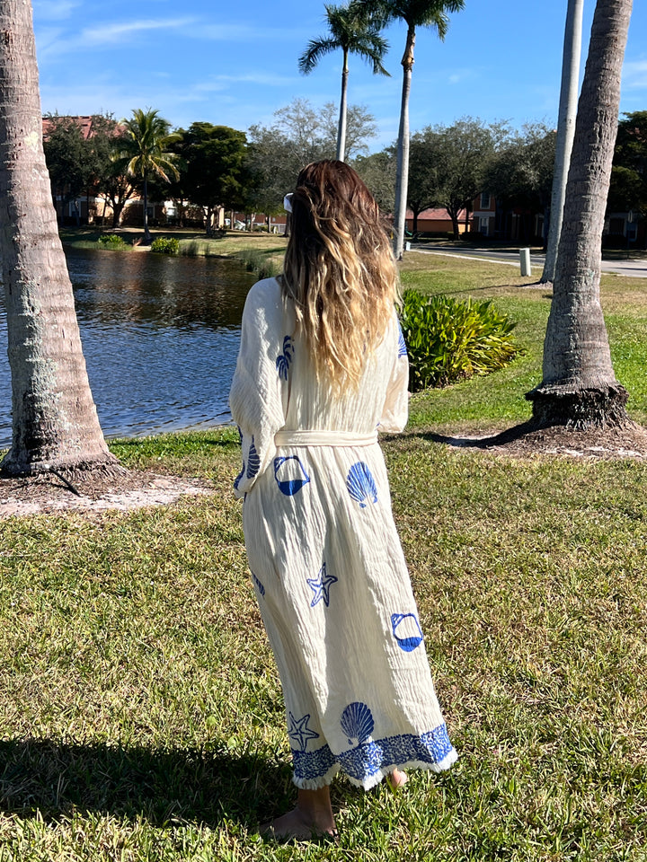 A woman with long, wavy hair stands on grass by a lake, wearing the Sea Shell Kimono Robe-Blue from turkanhome.com. The cream kimono features blue nautical prints. Palm trees and houses are visible in the background under a clear sky.