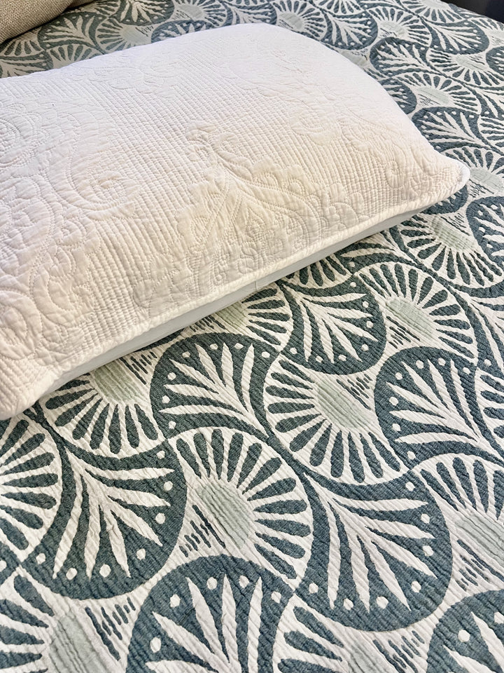 A white pillow with an embossed floral pattern sits atop a green and white geometric bedspread, paired with the Gingko Muslin Bed Blanket – Teal (King-Oversized Queen) from turkanhome.com for extra comfort.