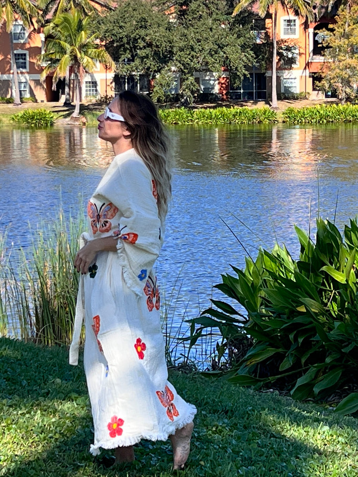A woman in the Butterfly Kimono Robe-Natural Color by turkanhome.com, featuring hand-printed butterfly and flower designs, stands barefoot on grass by a lake with palm trees and buildings in the background.