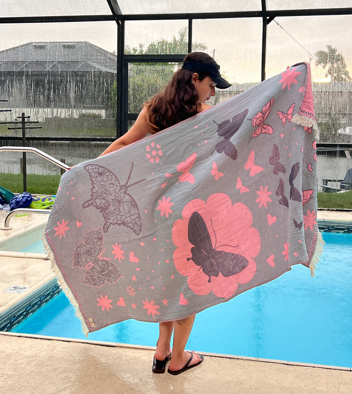 A woman in a black cap and flip-flops stands by a pool, holding the turkanhome.com Reversible Butterfly Towel—a quick-drying towel featuring pink and black butterflies and flowers. The enclosed pool area looks rainy outside.