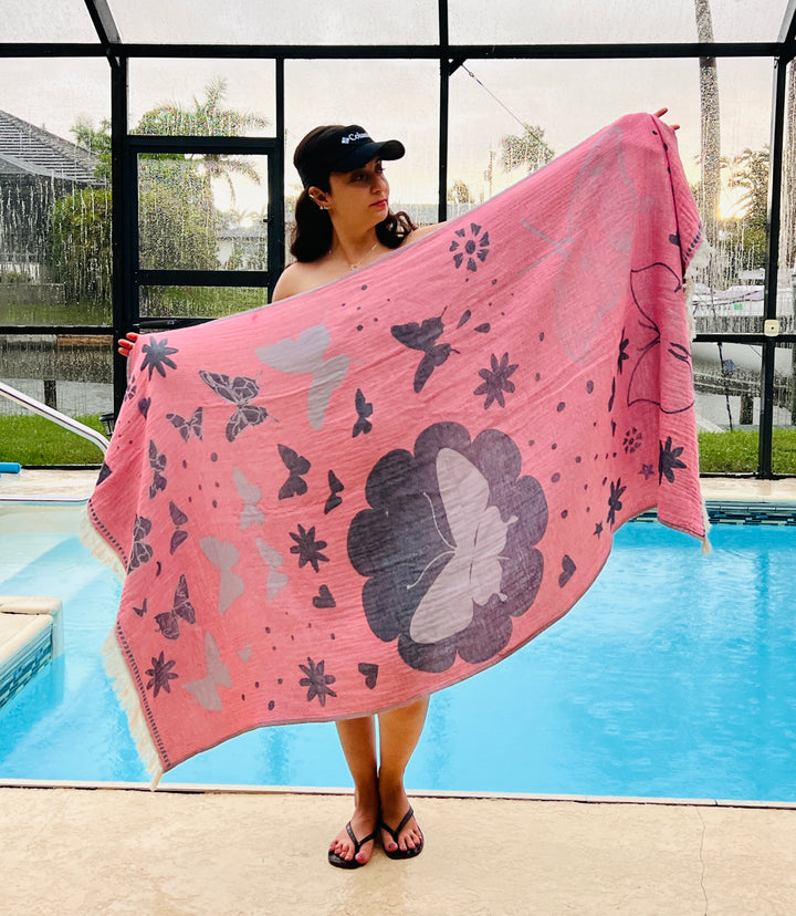 A woman in flip-flops and a black cap stands by the pool, showing off the turkanhome.com Reversible Butterfly Towel – Quick Drying Towel, featuring vibrant butterfly and flower prints. Palm trees and a screened patio complete the summer scene.