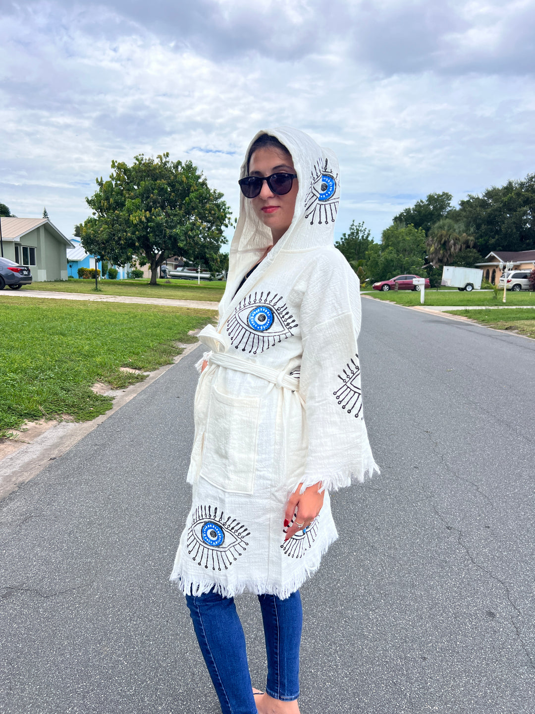 A barefoot person wears the Blue Eye With Lashes Kimono Robe (Short) by turkanhome.com, featuring eye designs, and stands on a suburban street with houses, lawns, and trees beneath a cloudy sky.