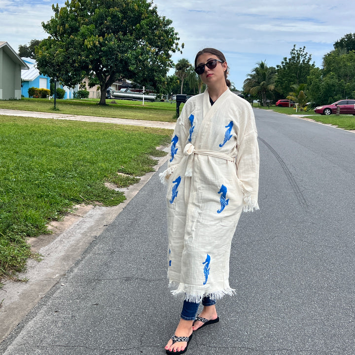 A woman in sunglasses models the Seahorse Kimono Robe from turkanhome.com, featuring a cream base with blue seahorse prints, as she poses casually on a suburban street surrounded by houses and greenery.