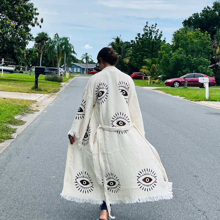 A person wearing the Brown Eye Kimono Robe by turkanhome.com walks barefoot down a tree-lined suburban street with grass and parked cars under a partly cloudy sky.