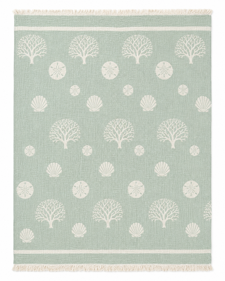 Infuse your space with a fresh coastal vibe by pairing this light green rug, adorned with white coral, sand dollar, and seashell patterns and finished with simple white stripes and fringed edges, alongside the Coastal Muslin Throw Blanket – Reversible Turkish Cotton from turkanhome.com.