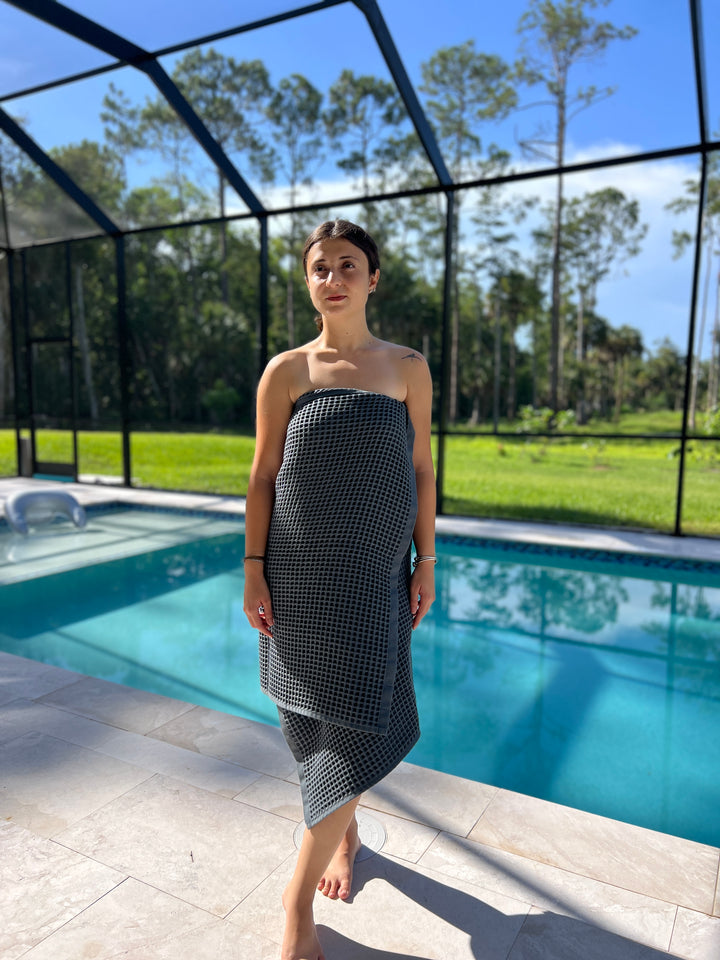 A woman by a pool wraps herself in a gray Waffle Bath Towel from turkanhome.com, with green grass and tall trees visible through a screened enclosure as sunlight casts gentle shadows on the deck.