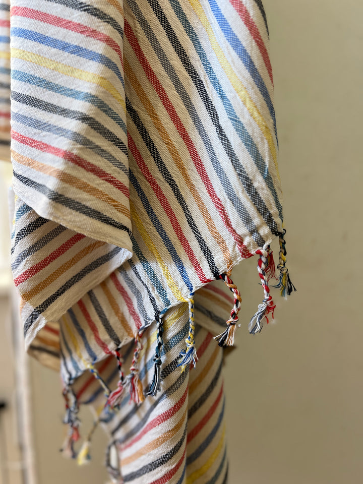 A close-up of the Mardin Handwoven Turkish Towel, Throw, Shawl by turkanhome.com shows diagonal yellow, blue, red, and white stripes with braided fringe on all-natural fibers and a softly blurred background.