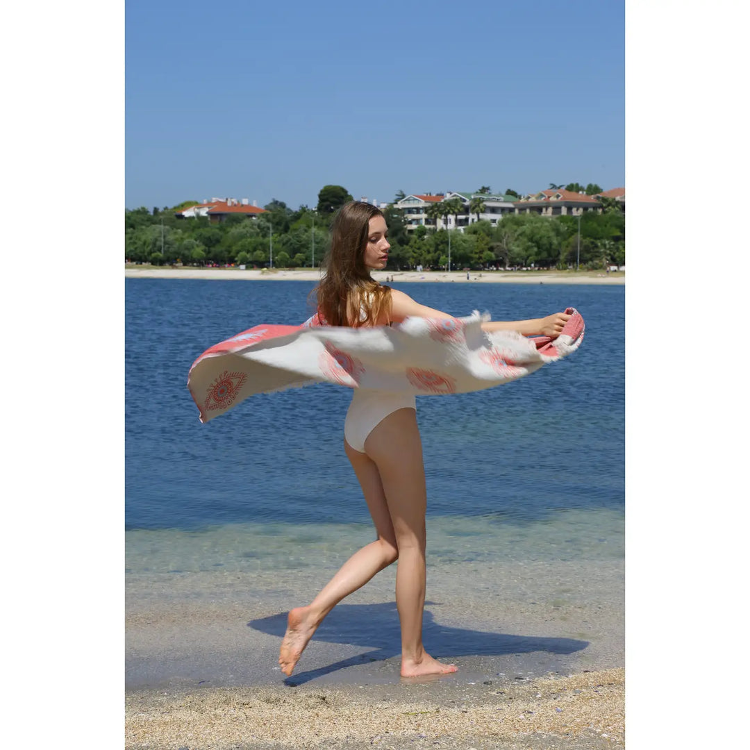 A woman in a white swimsuit stands on the sand by the water, holding the Eye Red Sand Resistant Turkish Beach Towel from turkanhome.com as it billows in the breeze. Trees and buildings appear in the background beneath a clear blue sky.