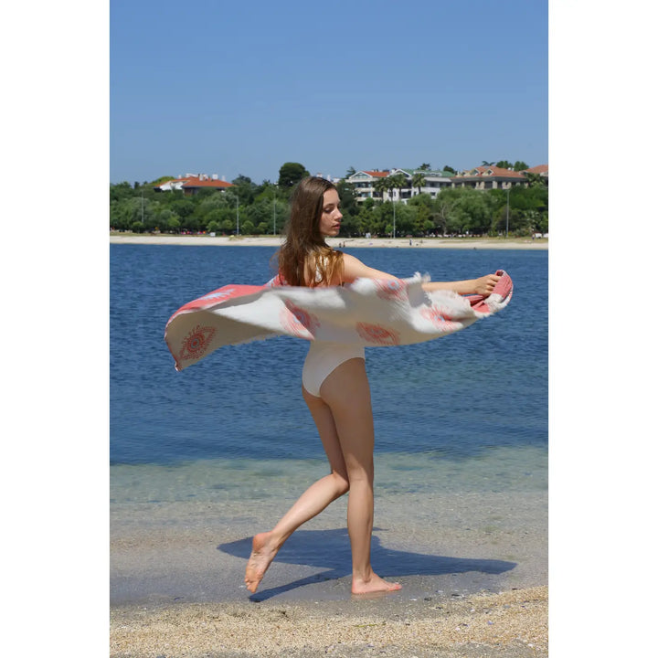 A woman in a white swimsuit stands on the sand by the water, holding the Eye Red Sand Resistant Turkish Beach Towel from turkanhome.com as it billows in the breeze. Trees and buildings appear in the background beneath a clear blue sky.