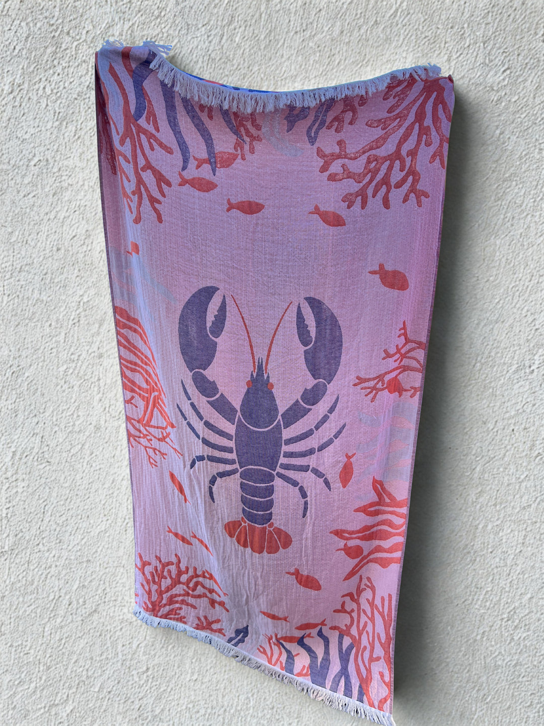 The Lobster Turkish Towel – Quick Drying Beach Towel by turkanhome.com, featuring a blue lobster with red coral and small fish on light pink fabric, hangs against a textured off-white wall.