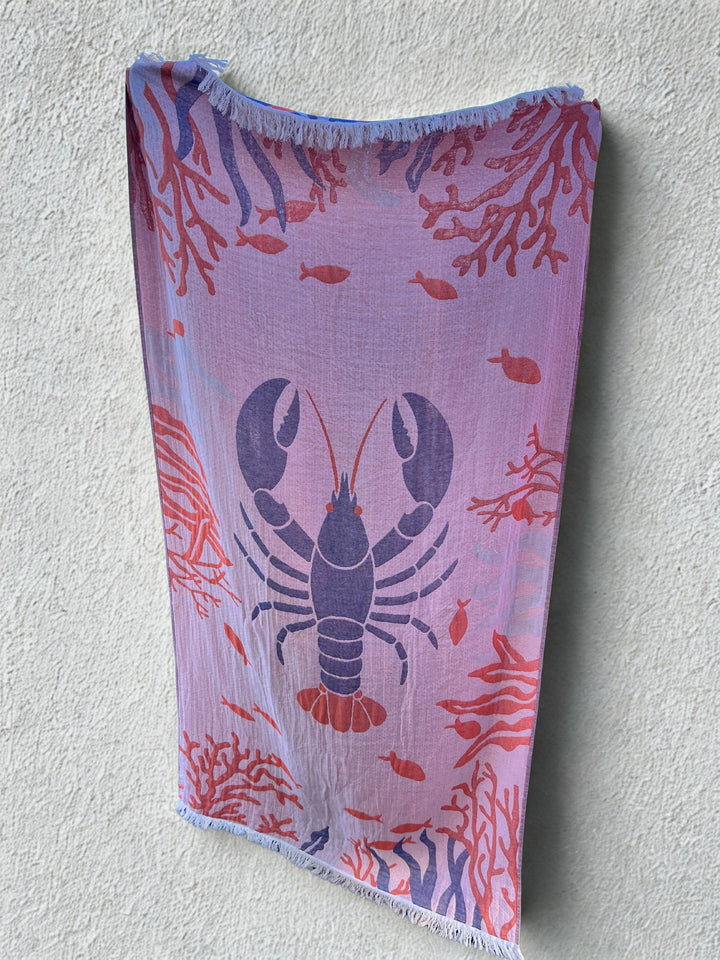 The Lobster Turkish Towel – Quick Drying Beach Towel by turkanhome.com, featuring a blue lobster with red coral and small fish on light pink fabric, hangs against a textured off-white wall.