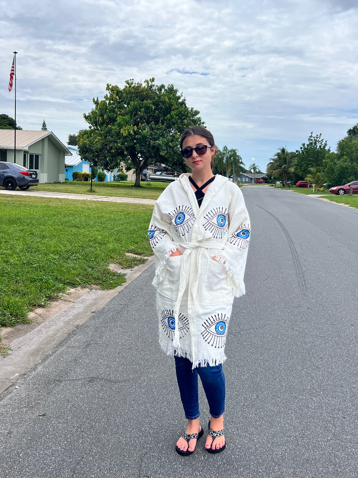A woman wearing sunglasses, jeans, sandals, and a Blue Eye With Lashes Kimono Robe by turkanhome.com stands on a suburban street lined with houses, trees, and an American flag under a cloudy sky.