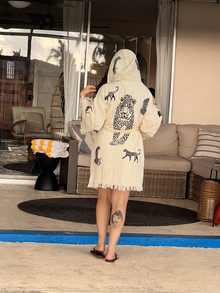 A person stands near a blue-edged step, wearing the Black Leopard Hooded Cotton Kimono Robe by turkanhome.com with their back to the camera, showing a leg tattoo. Outdoor patio furniture and a towel are in the background.