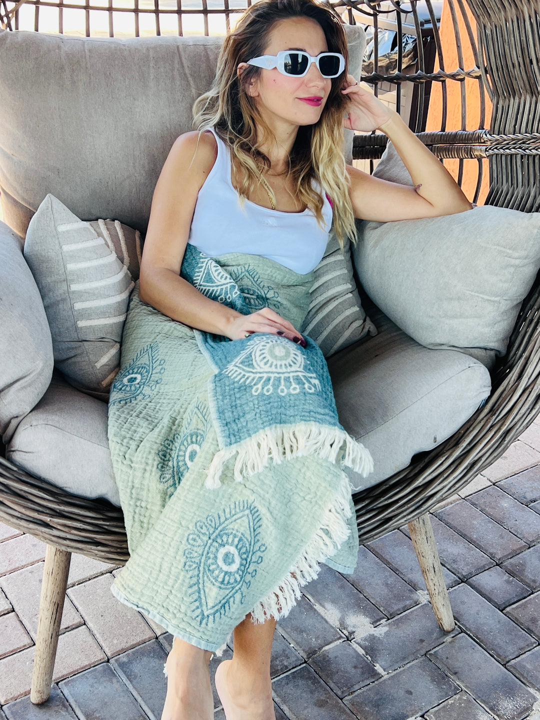 A woman wearing white sunglasses and a tank top lounges outdoors in a cushioned wicker chair, with the turkanhome.com Eye Sage Sand Resistant Turkish Beach Towel draped over her legs on a stone patio.