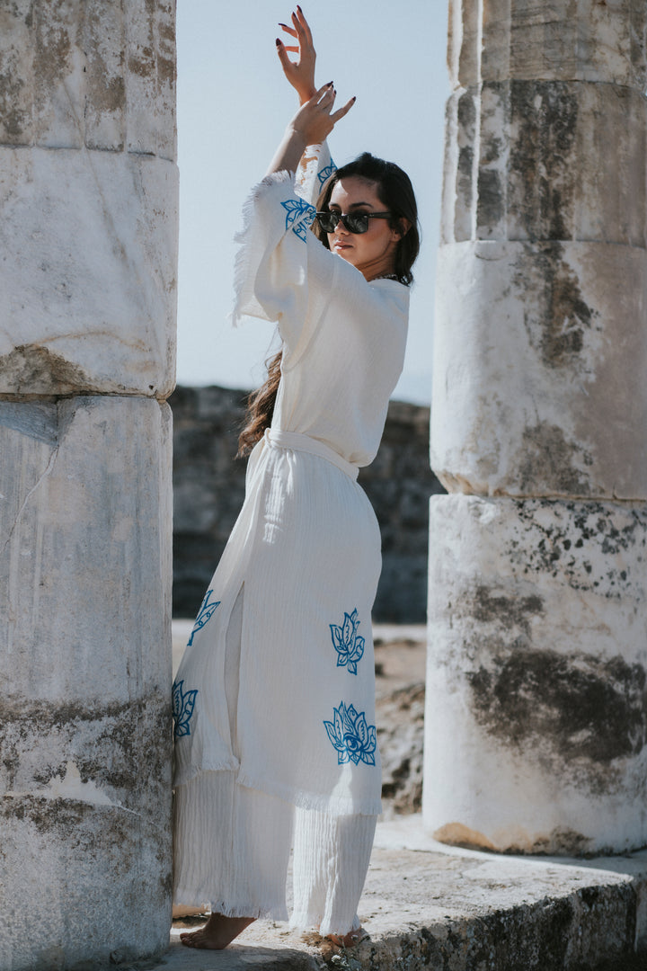 A woman in a Lotus Kimono Robe from turkanhome.com stands between ancient stone columns, sunglasses on and arms raised, as sunlight highlights the blue patterns and her festival boho kimono style.