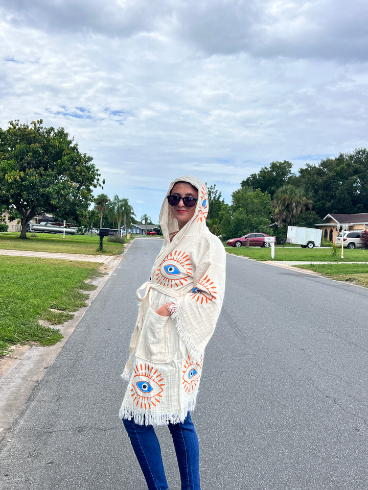 A person in the turkanhome.com Mystic Eye Kimono Robe Short with Pockets and Hood—made of natural cotton with colorful eye designs—stands on a cloudy suburban street with houses, cars, grass, and trees in the background.