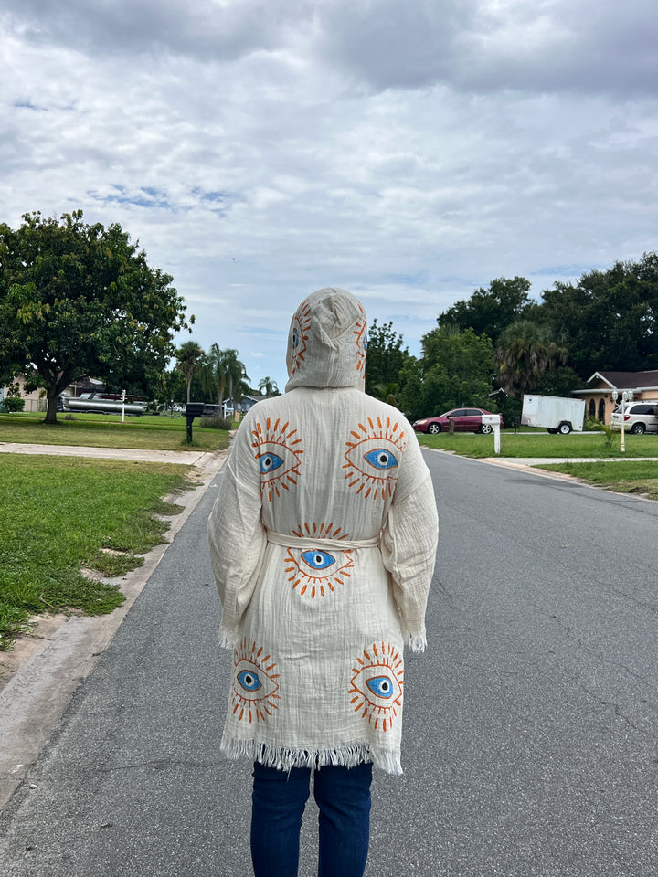 A person wears the turkanhome.com Mystic Eye Kimono Robe Short with Pockets and Hood—an off-white cotton robe with bold blue and orange eye designs—while standing on a cloudy suburban street among houses, trees, and cars.